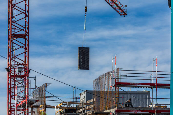 Ein Haus im Rohbau, das eingerüstet ist. Im linken Bildbereich befindet sich ein roten Kran,  welcher gerade Material an die Baustelle befördert.